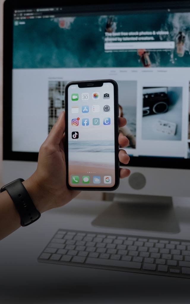Mão segurando celular com aplicativos de rede social em frente a um computador